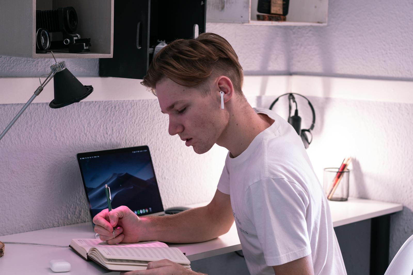Mladý muž se sluchátky píše poznámky u stolu, soustředěný na studium